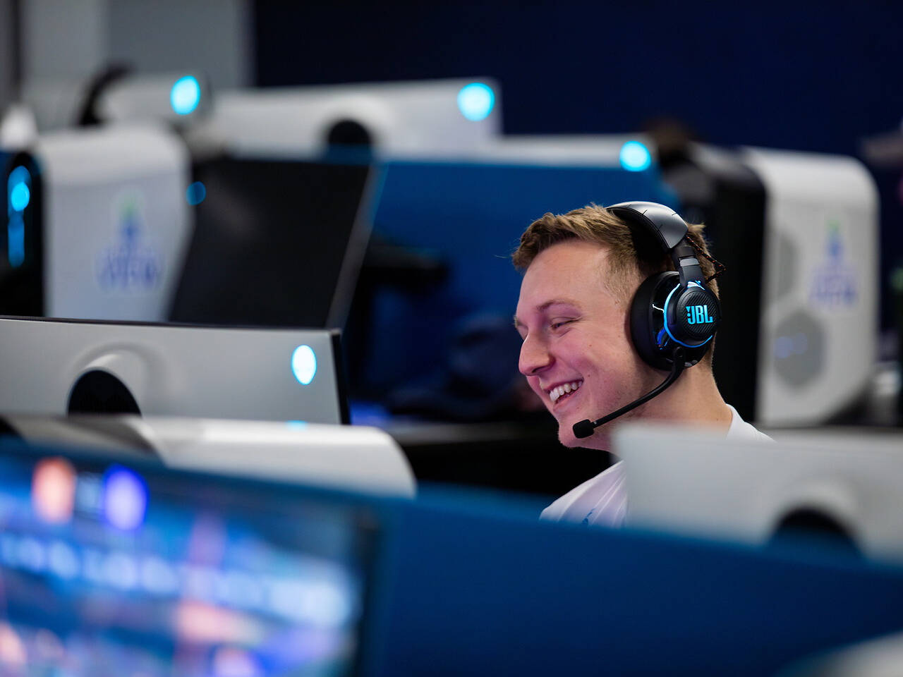 A person wearing a JBL headset smiles while seated at a computer station in a dimly lit room with multiple similar setups.
